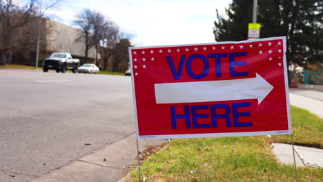 Vote-Here-Sign-Pointing-Right-with-People-Driving-Cars-in-Background,-Close-Up
