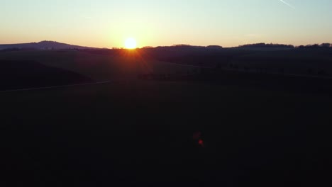 sun-rising-above-the-horizon-with-hills-behind-green-fields,-aerial,-crane-up