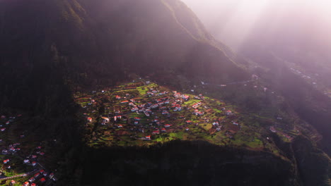 Cinematic-aerial-shot-of-Small-village-located-on-rural-hill-during-sunbeam-and-mountains-in-background