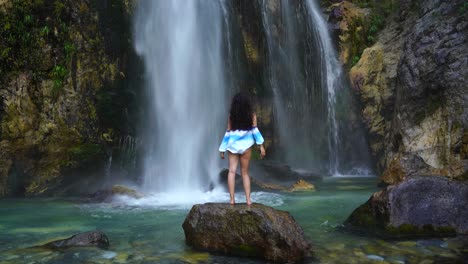 Girl-wonders-by-beautiful-waterfall,-standing-over-cliff-in-front-of-water-droplets-falling