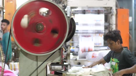 Male-Pakistani-Worker-Sorting-Plastic-Bags-At-Desk-With-Large-Spinning-Belt-Pully-Wheel-In-Factory-In-Karachi