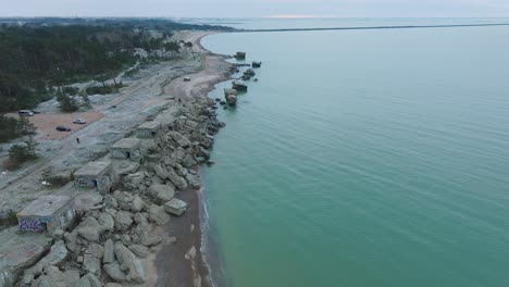 Establishing-aerial-view-of-abandoned-seaside-fortification-buildings-at-Karosta-Northern-Forts-on-the-beach-of-Baltic-sea-,-waves-splash,-overcast-day,-wide-drone-shot-moving-forward