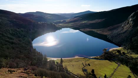 A-drone-shot-into-the-sun-of-The-Guinness-Lake-Lough-Tay-in-the-Wicklow-Mountains-Ireland-with-the-sun-reflecting-on-the-perfectly-still-calm-water-with-reflections-of-the-mountains-on-the-water