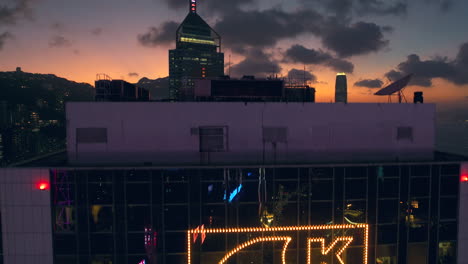 Aufsteigende-Luftaufnahme-Des-Zentralen-Platzes-Hinter-Dem-Sun-Hun-Kai-Gebäude-Und-Der-Skyline-Von-Hong-Kong-Im-Hintergrund-A-Während-Des-Goldenen-Sonnenuntergangs