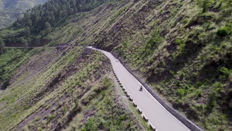 Aerial-drone-long-shot-of-a-man-riding-a-bike-with-luggage-traveling-on-an-empty-scenic-road-in-a-cold-mountainous-region-or-passing-by-lush-green-farms-and-a-valley