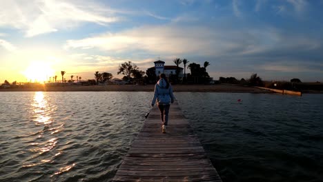Young-woman-walking-through-a-wooden-pier-bridge-by-the-beach-at-sunset