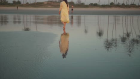 Woman-in-a-yellow-dress-calmly-walking-barefoot-and-swinging-her-arms-at-the-shoreline-at-dawn