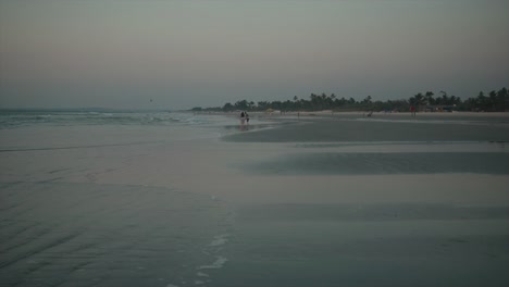 Playa-Tranquila-Con-Pequeñas-Olas-Aplastando-Tranquilamente-Contra-La-Orilla-Y-Dos-Personas-Jugando-En-El-Agua-A-Distancia