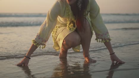 Frau-In-Einem-Hellgelben-Kleid-Und-Braunen-Armbändern-An-Beiden-Händen,-Die-Mit-Ihren-Händen-Im-Sand-Am-Ufer-Hockt