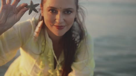 Woman-in-a-light-yellow-dress-playing-with-a-starfish-at-the-beach-with-waves-in-the-background