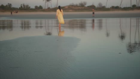 Woman-in-a-yellow-dress-calmly-walking-at-a-distance-at-the-shoreline-at-dawn