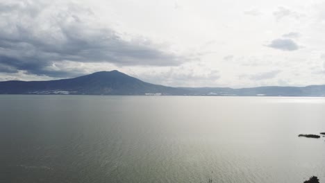 360-turnaround-drone-aerial-shot-of-a-huge-lake-near-a-small-town-with-mountains-in-the-background-and-clouds,-60FPS-video