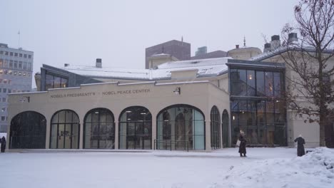 Snow-Falling-Outside-The-Nobel-Peace-Center-In-Oslo,-Norway-With-Two-People-Outside-And-Then-Walking-Past