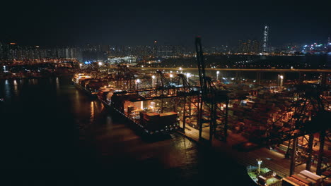 Container-vessels-moored-under-flood-lights-at-a-terminal-with-skyline-of-Hong-Kong-in-backdrop-Drone-night-panning-shot