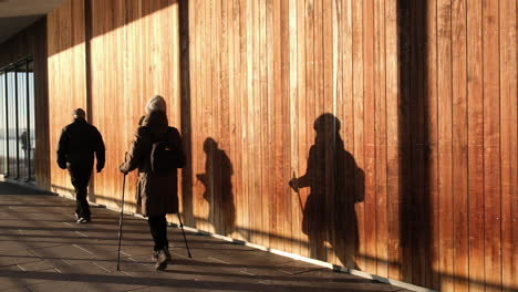 Leute-In-Mütze-Und-Jacke,-Die-Auf-Einem-Kalten-Sonnenuntergang-In-Oslo,-Norwegen,-Im-Flur-Eines-Sonnenbeschienenen-Gebäudes-Spazieren