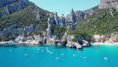 Tourist-Boats-Relax-at-Turquoise-Blue-Sea-at-Cala-Goloritze-Beach,-Sardinia,-Italy---Aerial-4k-Circling