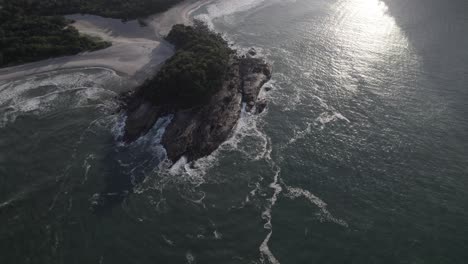 Drone-above-the-sea-showing-a-rock-island-and-the-atlantic-rainforest-behind-at-the-light-of-the-morning,-with-fog-in-a-sunny-day