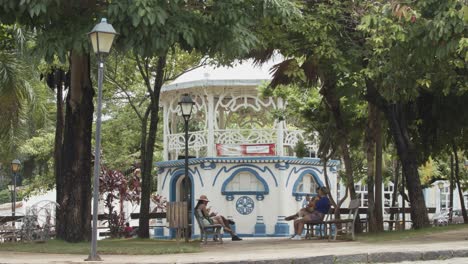 Gente-En-Los-Bancos-Del-Parque-Junto-A-Un-Quiosco-De-Música-En-Un-Parque-De-La-Ciudad