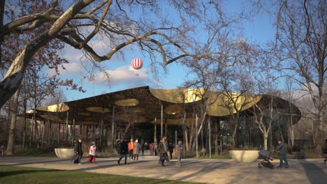 The-House-of-Music,-Hungary---designed-by-Sou-Fujimoto--at-Budapest-City-Park-on-a-sunny-winter-afternoon-with-a-huge-sightseeing-balloon-in-the-background---January-2023