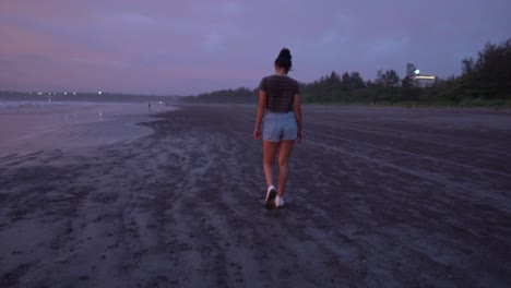 Young-lady-in-grey-shirt,blue-jeans-and-white-shoes-walking-on-the-beach-with-city-lights-in-the-background