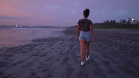 Girl-in-blue-shorts-walking-at-a-beach-with-calm-waves-touching-the-shore-at-sunset