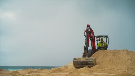 An-orange-construction-excavator-scoops-and-drops-sand-on-a-beach-by-the-ocean,-mid-shot