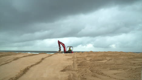 An-orange-construction-excavator-moves-up-and-down-the-beach-smoothing-out-the-sand,-Sydney-Australia