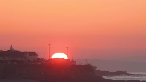 Sun-Rising-timelapse-From-Behind-Downtown-Estoril-with-some-cars-driving-on-the-coastal-road,-Portugal