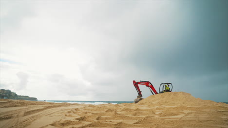 An-orange-construction-excavator-scoops-and-drops-sand-on-a-beach-by-the-ocean,-in-Sydney,-Australia