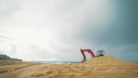 Una-Excavadora-De-Construcción-Naranja-Recoge-Y-Deja-Caer-Arena-En-Una-Playa-Junto-Al-Océano,-En-Sydney,-Australia