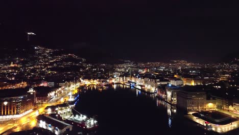 Aerial-dolly-forward-over-city-fjord-towards-bright-night-lights-in-Bergen---Norway