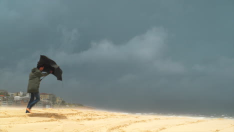 A-man-walks-along-a-sandy-beach-holding-an-umbrella-on-a-stormy-and-windy-day,-Slow-motion