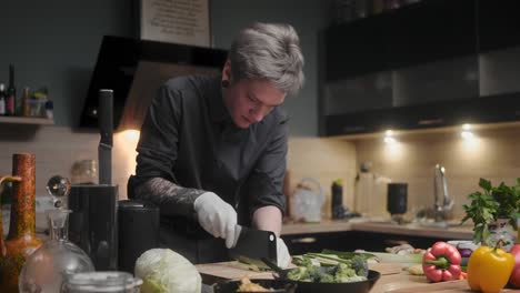 Young-professional-male-chef-in-an-elegant-black-shirt-with-an-alternative-look,-tattoos,-gloves-slicing-fresh-zucchini-A-modern,-fancy-looking-kitchen-with-fresh-vegetables-besides-Cinematic-Sliding