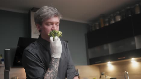 Young-crazy-male-chef-in-an-elegant-black-shirt-with-an-alternative-look,-tattoos-smelling-fresh-broccoli-rolling-eyes-in-lust-A-modern,-fancy-looking-kitchen-with-fresh-vegetables-besides-Cinematic