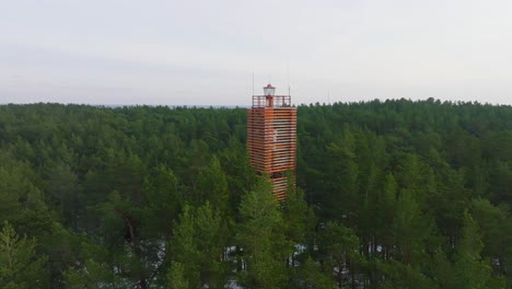 Aerial-view-of-Bernati-lighthouse-surrounded-by-lush-green-pine-tree-forest-with-light-snow,-Nordic-woodland,-Baltic-sea-coast,-sunny-winter-day,-Latvia,-wide-ascending-drone-shot-moving-forward