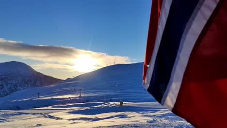 Norwegian-flag-in-foreground-with-alpine-skiing-slopes-and-sun-in-background---Handheld-clip-Myrkdalen-Norway