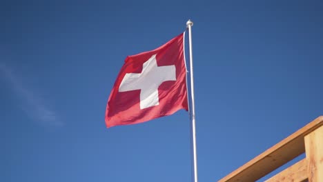 Bandera-Suiza-Soplando-Y-Ondeando-En-El-Viento-Unida-A-Un-Poste-Blanco-En-Un-Día-Soleado-Con-Un-Esquí-Azul,-Cámara-Lenta-4k