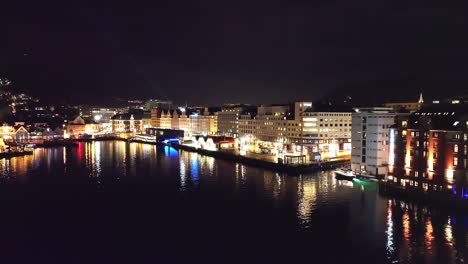 Antena-Nocturna-Que-Muestra-El-Mercado-De-Pescado-De-Bergen-Y-El-Centro-De-La-Ciudad-Alrededor-De-Strandkaien---Ciudad-Iluminada-Con-Reflejos-En-Un-Fiordo-Tranquilo