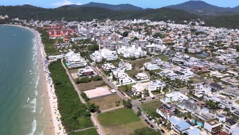 Escena-De-Altitud-De-Drones-Aéreos-De-Playa-Turística-Con-Muchos-Alojamientos-Frente-Al-Mar,-Edificios-De-Vuelo-Y-Casas-Con-Gente-Divirtiéndose-En-La-Playa-En-Florinópolis-Jurere-Internacional