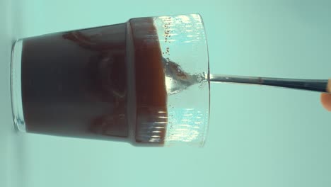 A-vertical-close-up-shot-of-a-glass-of-black-coffee-stirring-wih-a-silver-spoon,-floating-cafe-grains,-studio-light,-4K-video