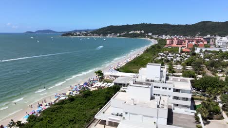 Aerial-drone-scene-of-tourist-beach-with-many-accommodations-facing-the-sea,-flight-close-to-buildings-and-houses-with-blue-sea-and-people-having-fun-on-the-beach-in-Florinópolis-Jurere-Internacional