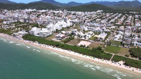Luftdrohne-Höhenszene-Eines-Touristischen-Strandes-Mit-Vielen-Unterkünften-Mit-Blick-Auf-Das-Meer,-Fluggebäude-Und-Häuser-Mit-Menschen,-Die-Sich-Am-Strand-In-Florinópolis-Jurere-Internacional-Vergnügen