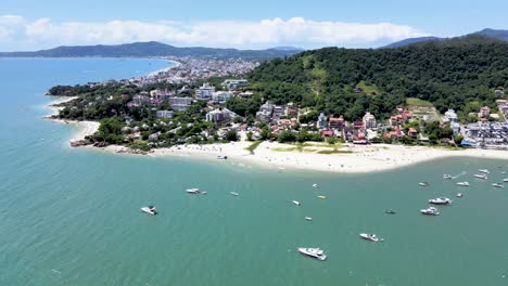 Aerial-drone-scene-of-paradise-beach-with-many-boats-and-yachts-in-the-sea-with-pier-and-buildings-facing-the-sea-in-Canajure-beach-Florianopolis-Santa-Catarina