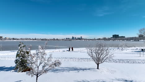 Drone-moving-past-snowy-trees-at-Sloan-Lake-during-winter-storm,-revealing-a-family-walking-with-Denver-City-in-the-background
