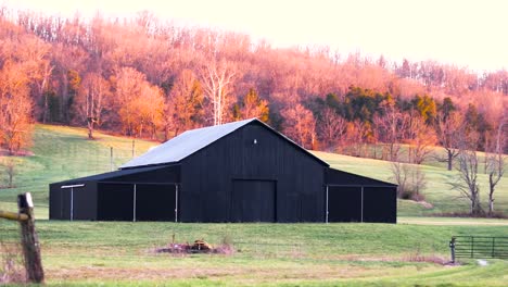 Still-image-of-a-black-barn-at-the-base-of-a-hillside-covered-with-fall-foliage-in-Kentucky