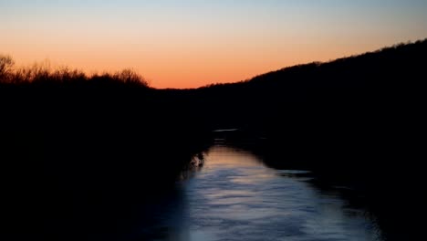 Beautiful-sunset-on-stream,-lined-by-shadowy-brush-at-dusk