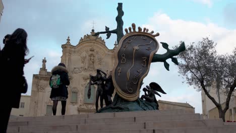 Clock-sculpture-in-Matera,-Italy-with-people-walking-around