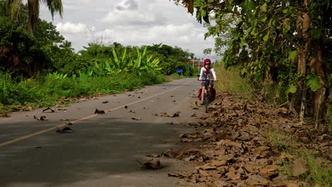 Asian-female-slowly-biking-through-leaves-on-paved-road-towards-camera,-Thailand