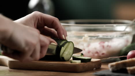 Close-up-of-amateur-cook-cutting-fresh-cucumber-into-small-chunks-to-prepare-vegetable-spread-on-wooden-kitchen-table