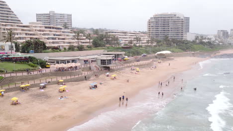 Drone-Aéreo-Disparado-A-Lo-Largo-De-La-Playa-Principal-De-Umhlanga-Con-Personas-Sentadas-Bajo-Sombrillas